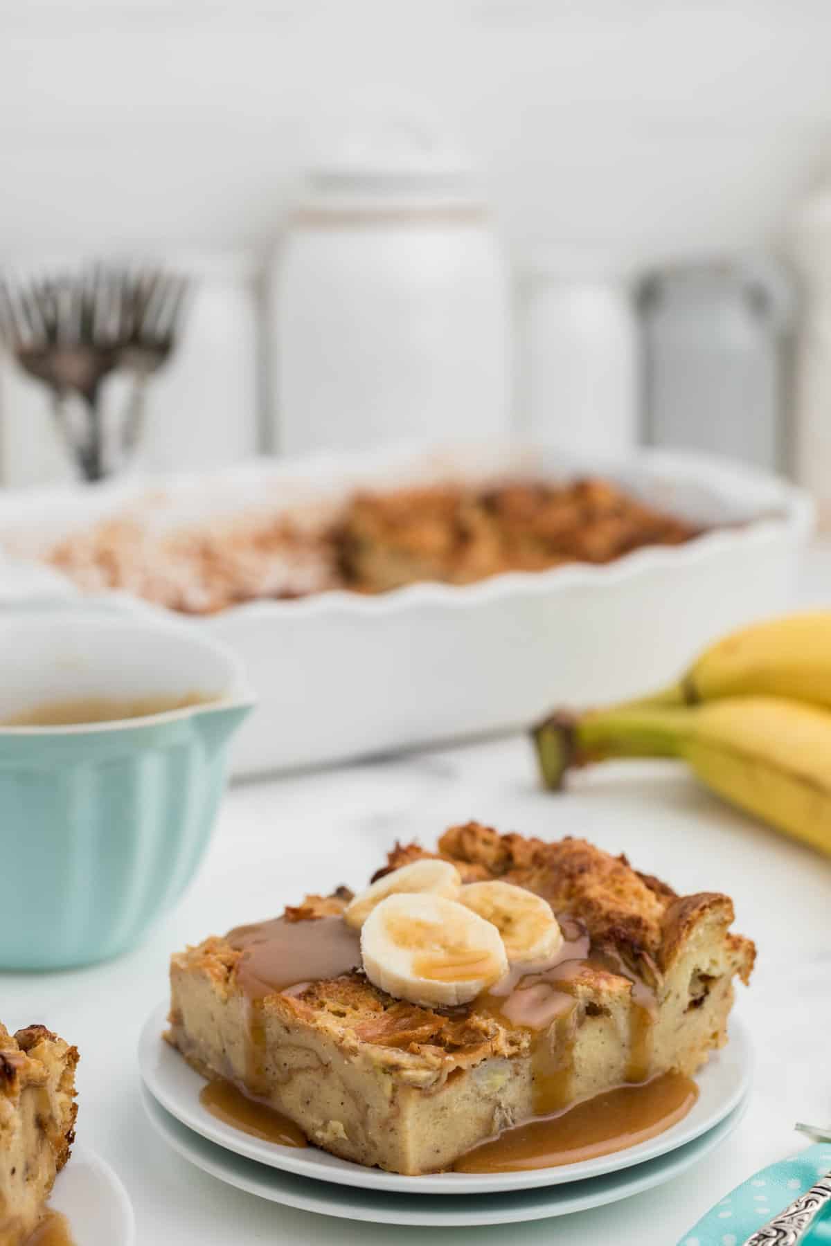 A plate with a slice of bread pudding is topped with caramel and banana slices.