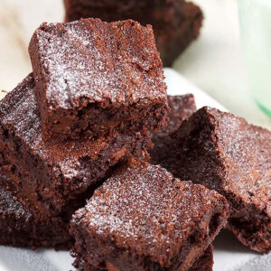 Brownies piled on a white plate with a glass of milk in the background.