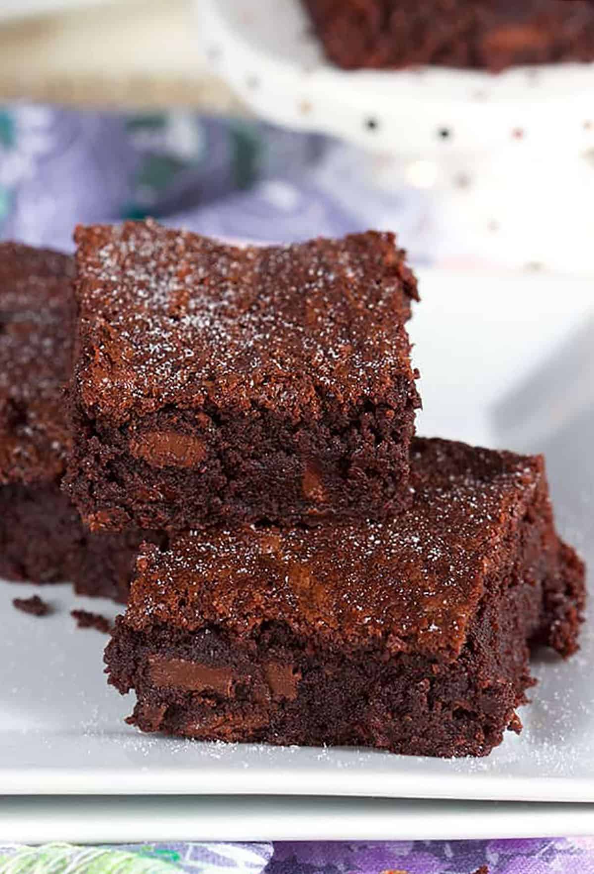 Three brownies piled on a white plate with confectioners sugar sprinkles on top.