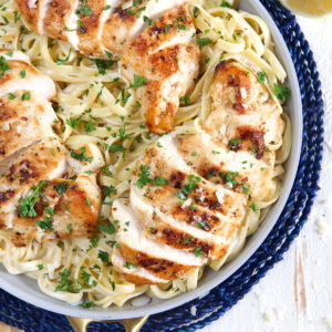 A pan of sliced chicken breast and creamy fettuccine is presented on a blue place mat.