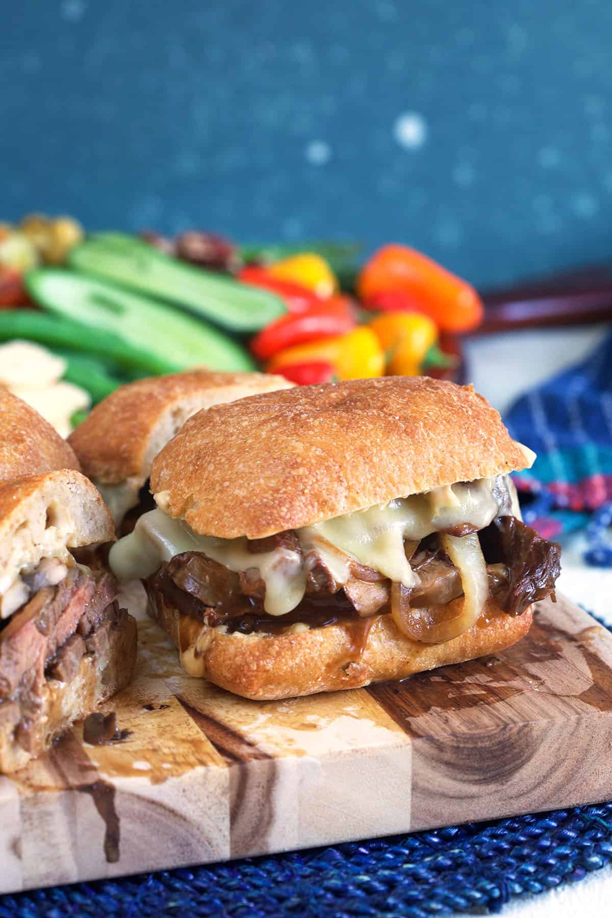 Several roast beef sandwiches are presented on a wooden cutting board next to some fresh vegetables.