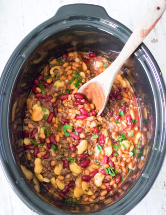 A slow cooker filled with cooked calico beans is being mixed with a wooden stirring spoon.