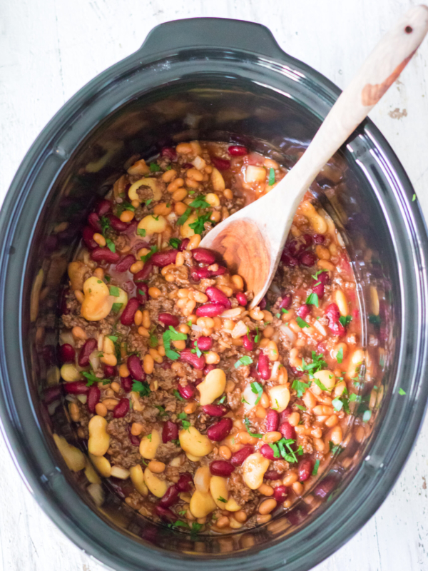 A slow cooker filled with cooked calico beans is being mixed with a wooden stirring spoon.