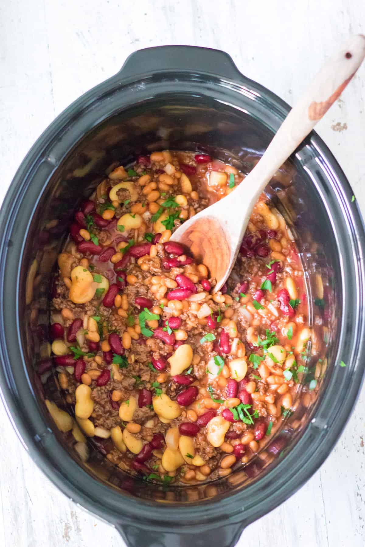 A slow cooker filled with cooked calico beans is being mixed with a wooden stirring spoon.