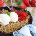 Skillet with a blue and white striped towel tied to the handle filled with strawberry rhubarb crisp with two wooden forks in it.