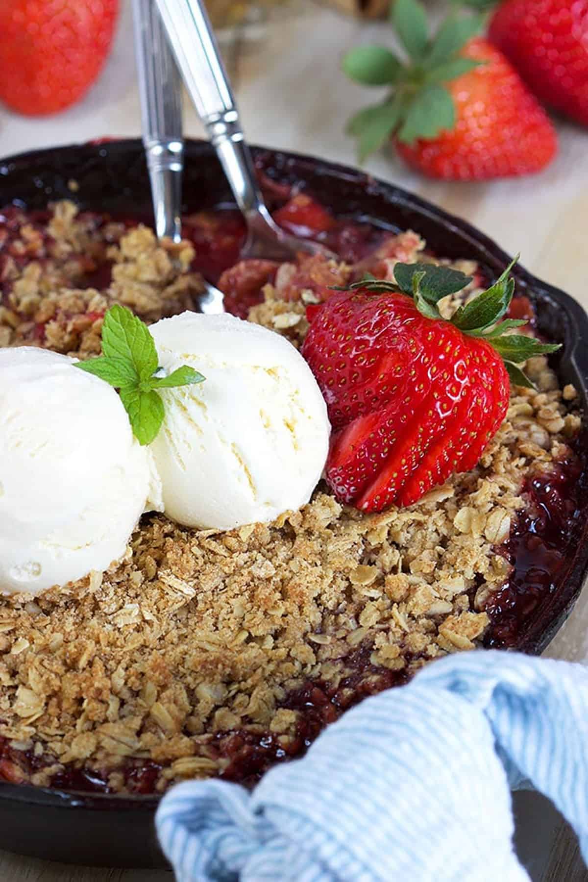 Strawberry Rhubarb Crisp in a skillet with a light blue towel tied to the handle. Two scoops of vanilla ice cream and a strawberry on top.