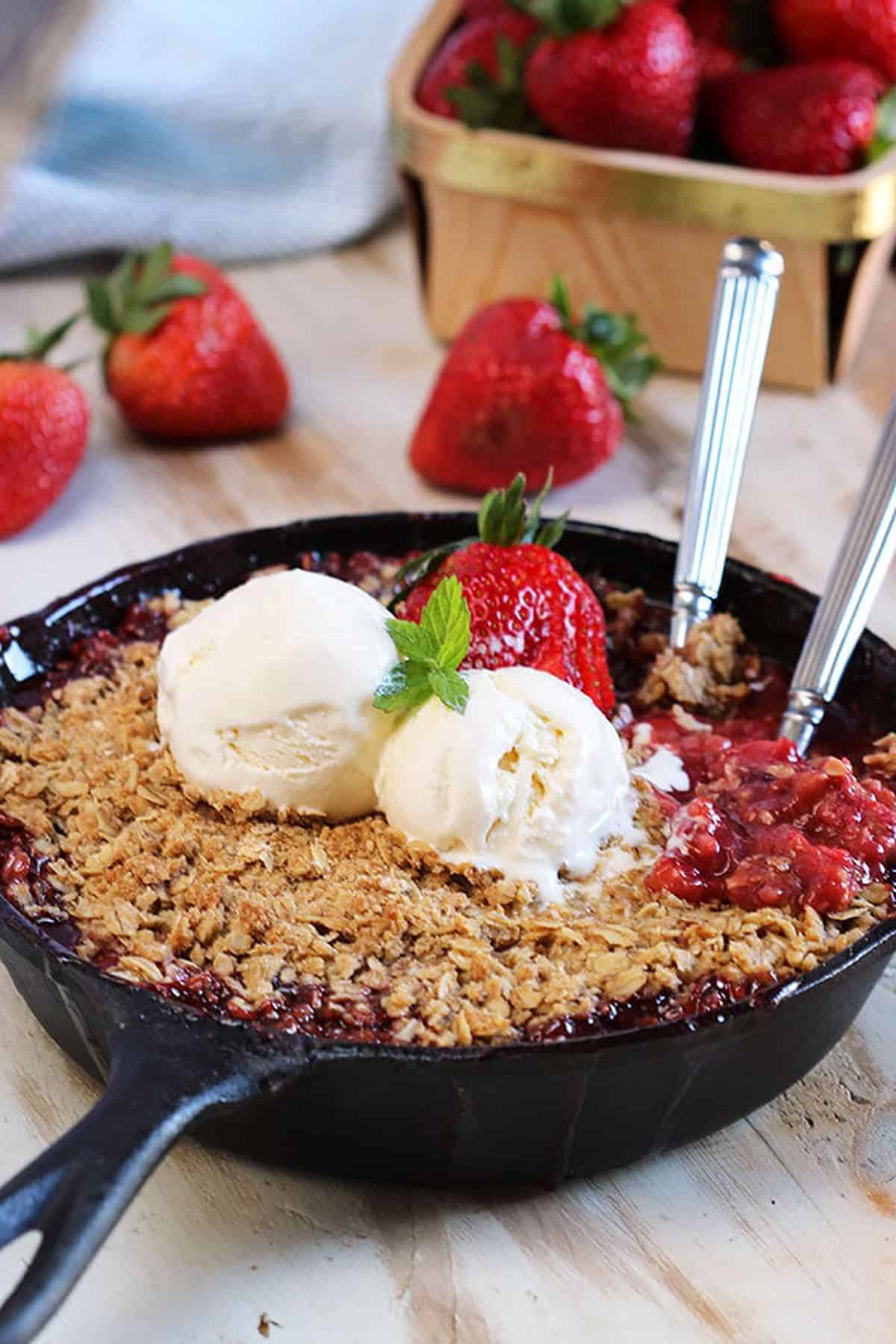 Strawberry Rhubarb Crisp in a cast iron skillet with two forks in it and two scoops of ice cream on top. Berries in a basket in the background.