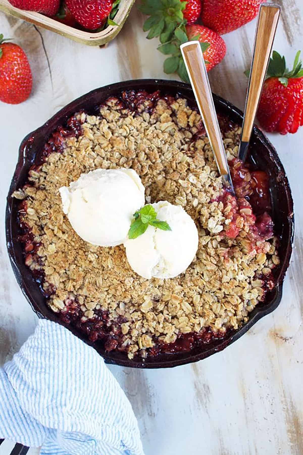 Cast iron skillet with strawberry rhubarb crisp and two scoops of vanilla ice cream and a blue and white striped towel tied to the handle.