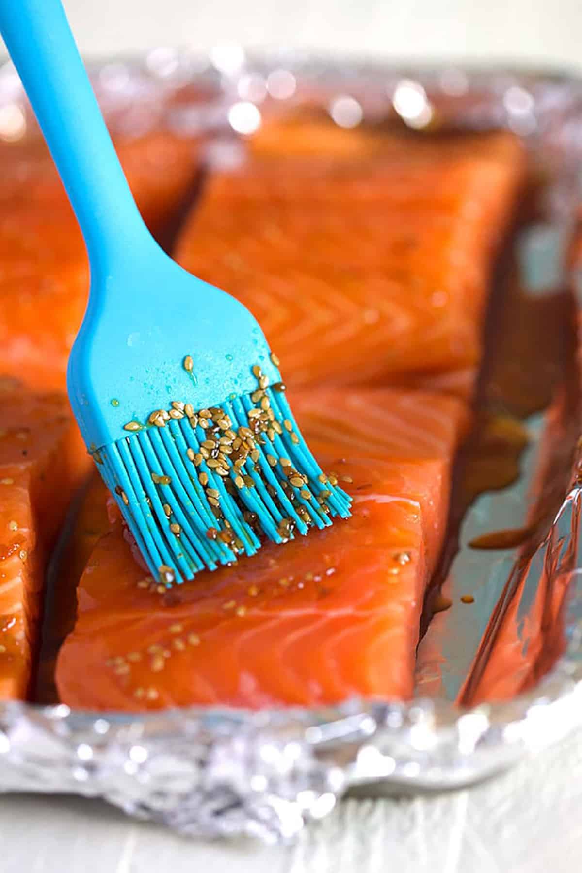 Blue basting brush applying teriyaki sauce to salmon filets on a baking sheet.