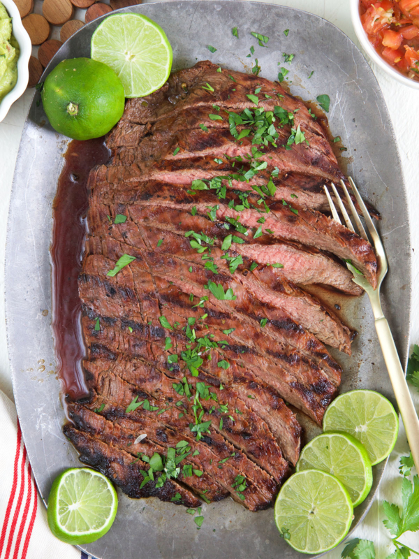 Carne asada has been sliced and is served on a gray serving platter with cilantro and limes.