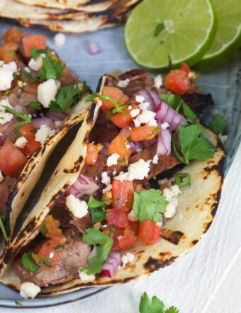 Two carne asada tacos are garnished with fresh cilantro, pico de gallo, and cojita cheese.