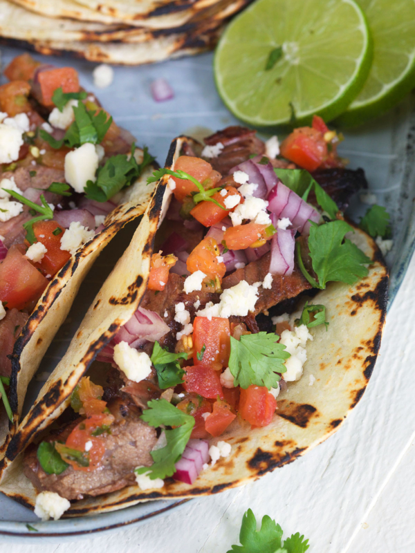 Two carne asada tacos are garnished with fresh cilantro, pico de gallo, and cojita cheese.