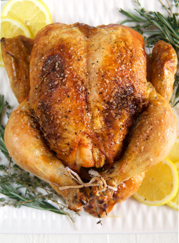 A fully cooked whole chicken is presented on a white serving tray with rosemary sprigs and lemon slices.
