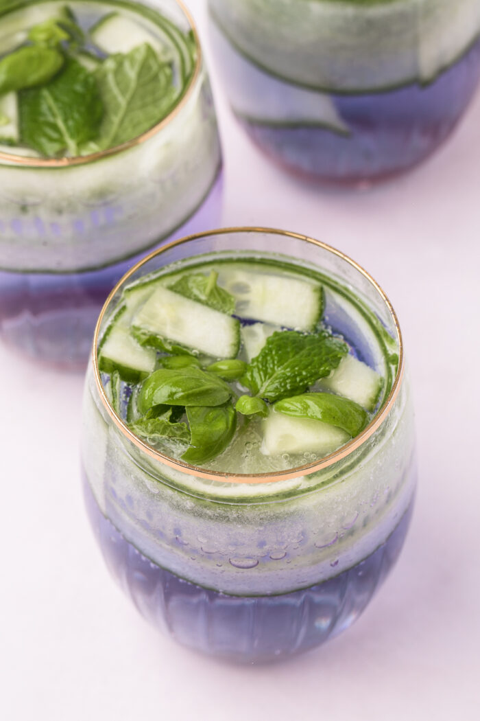 Three full glasses are garnished with limes, cucumber, and herbs.