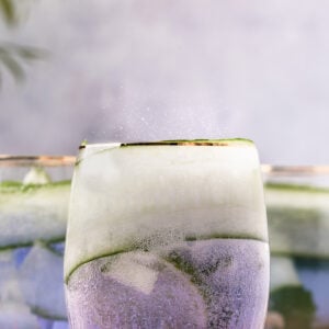 A glass is filled with blue, fizzy liquid and thin cucumber slices.