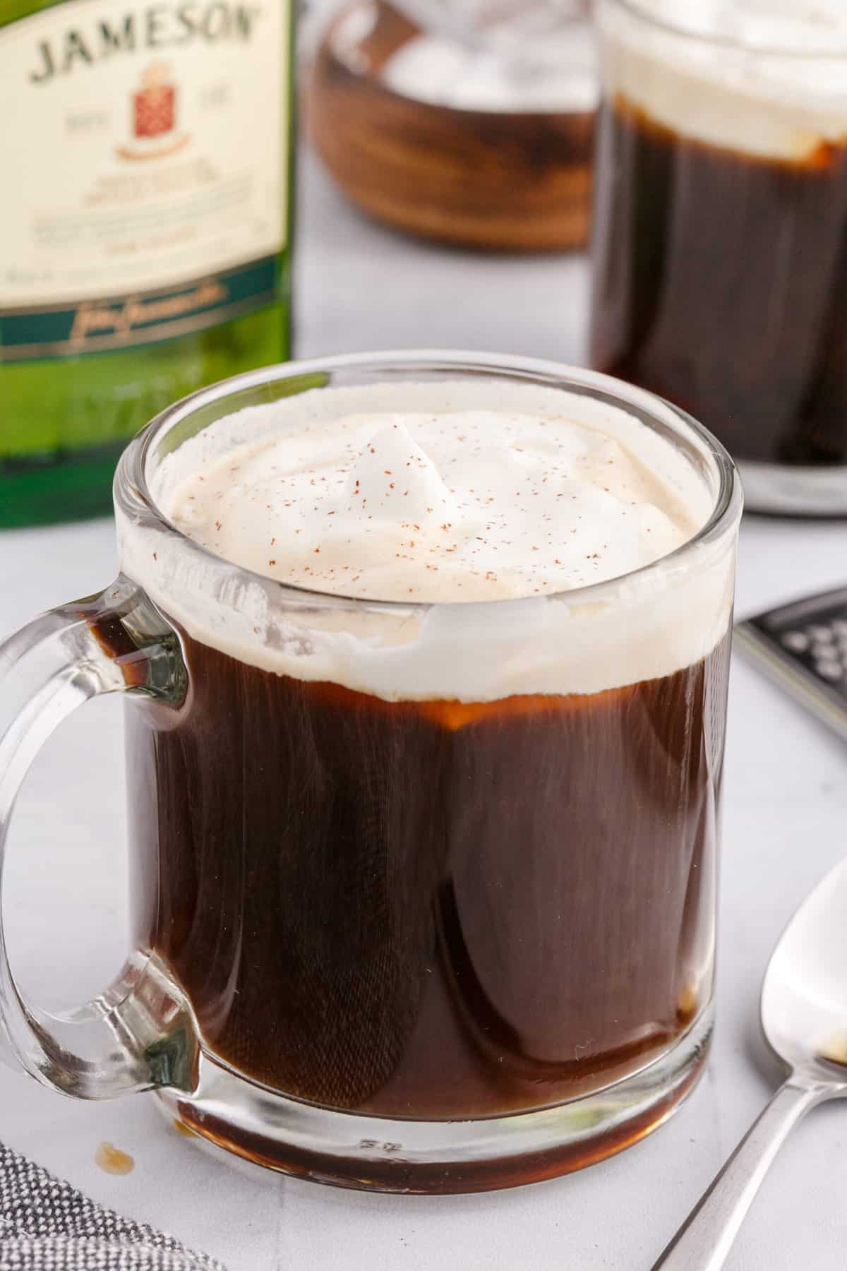 A glass mug filled with irish coffee and whipped cream is placed next to a spoon.
