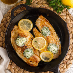 A black pan containe two cooked fish filets that are topped with lemon slices and garlic butter.