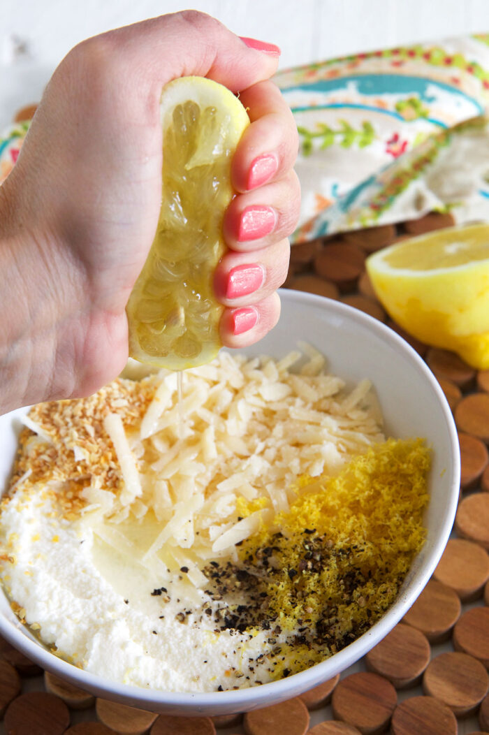 A lemon is being squeezed over a bowl filled with cheese, seasonings, and lemon zest.