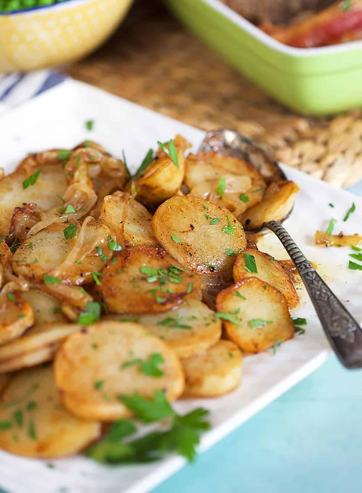 Lyonnaise potatoes on a white platter with a spoon and peas in the background.