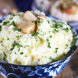 Roasted Garlic Mashed Potatoes with blue and white bowl with roasted garlic cloves on top.