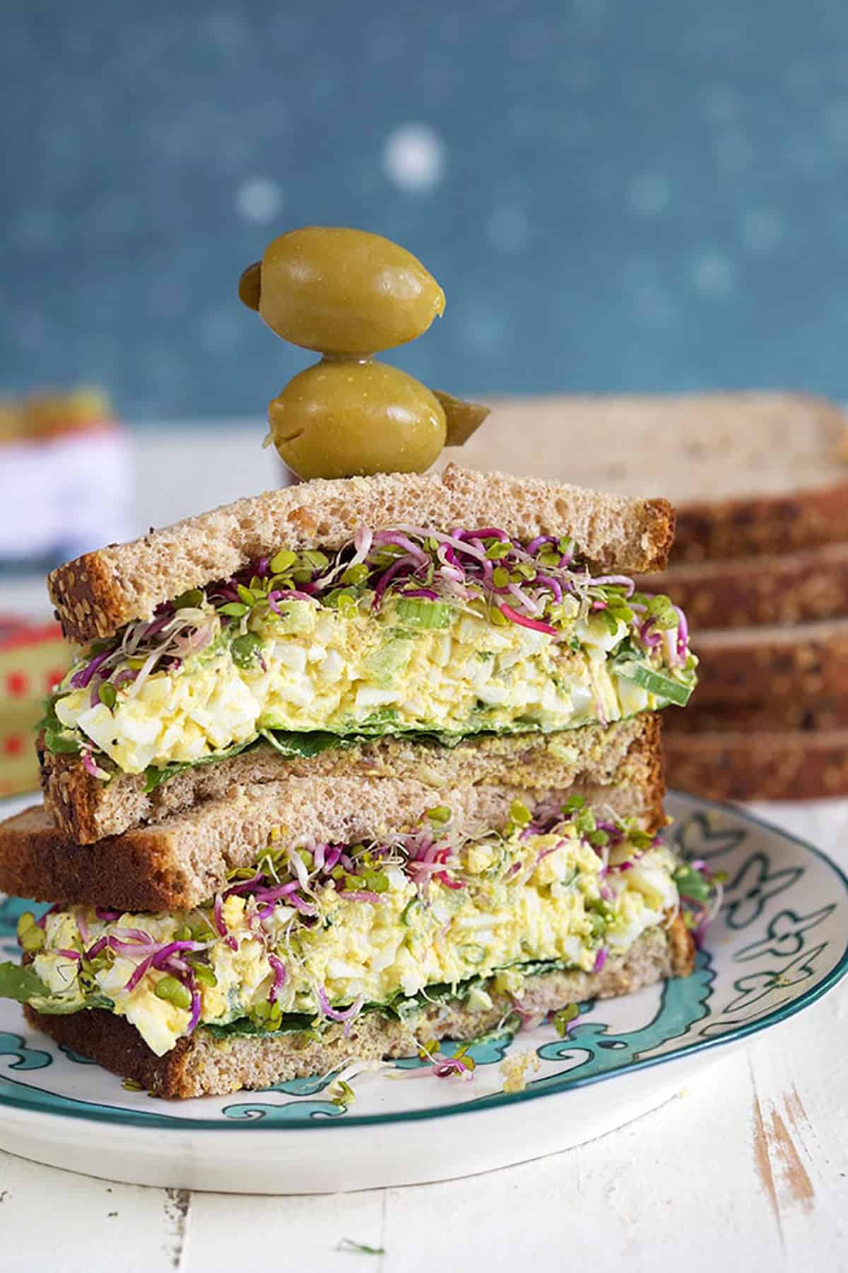 Egg salad sandwich with olives on top on a blue and white plate.