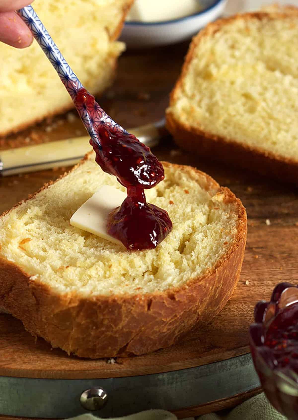 Slice of brioche bread with a pat of butter and a spoon dropping jam on top.
