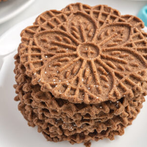 A stack of chocolate pizzelles are presented on a round white plate.