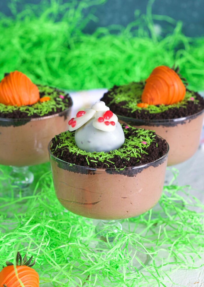 Three dirt pudding cups are presented on a white surface, surrounded by decorative green straw.