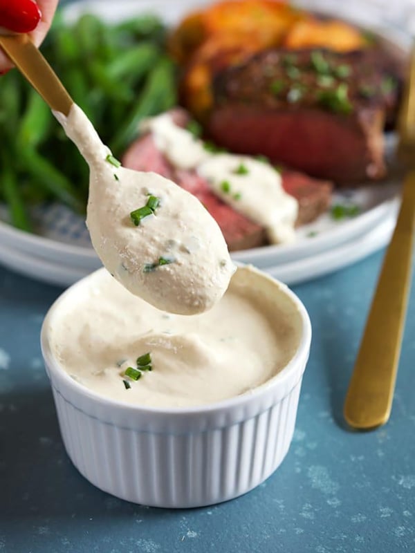 Creamy Horseradish Sauce on a gold spoon over a bowl of horseradish sauce on a blue background.