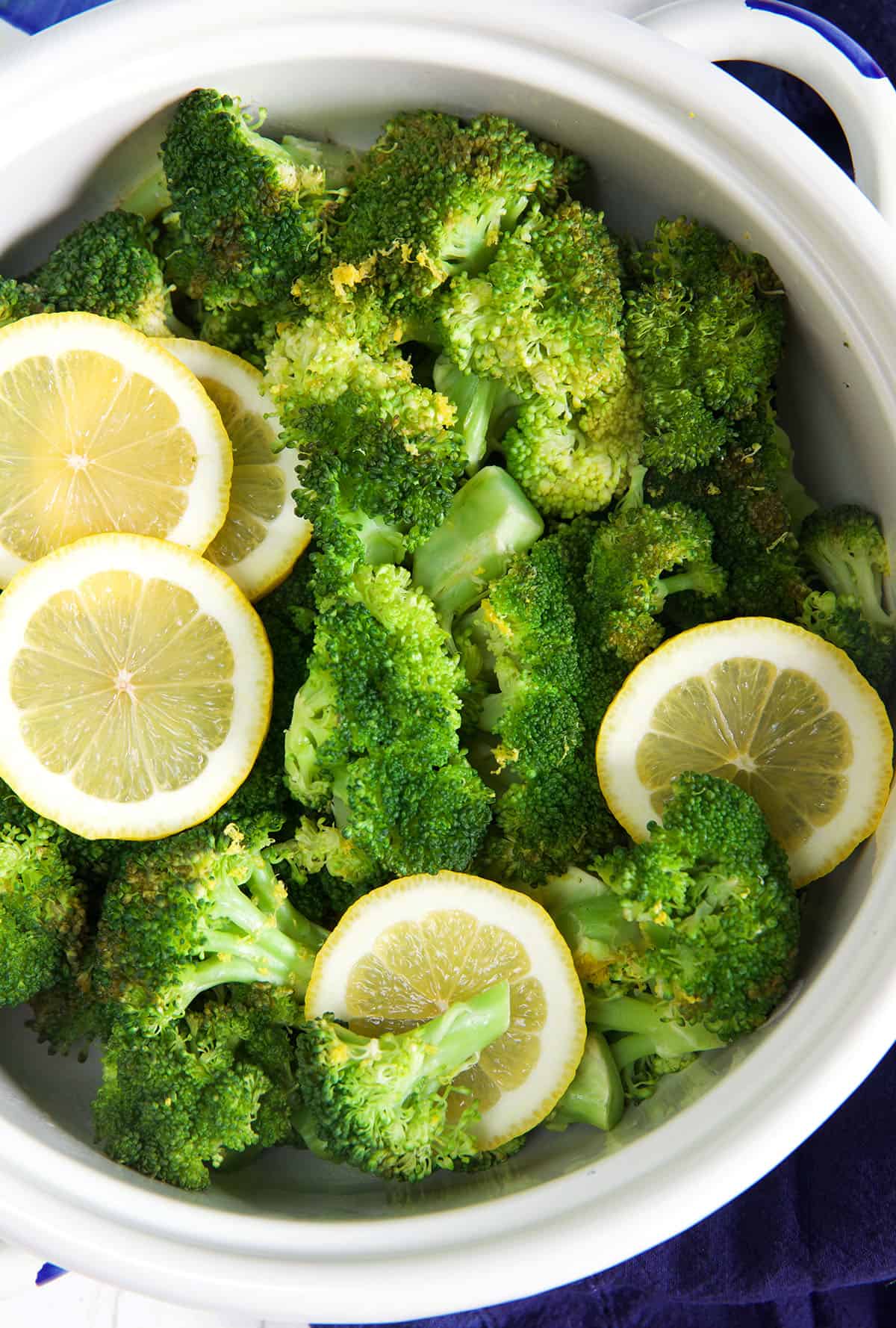 A white pot is filled with freshly steamed broccoli and lemons.