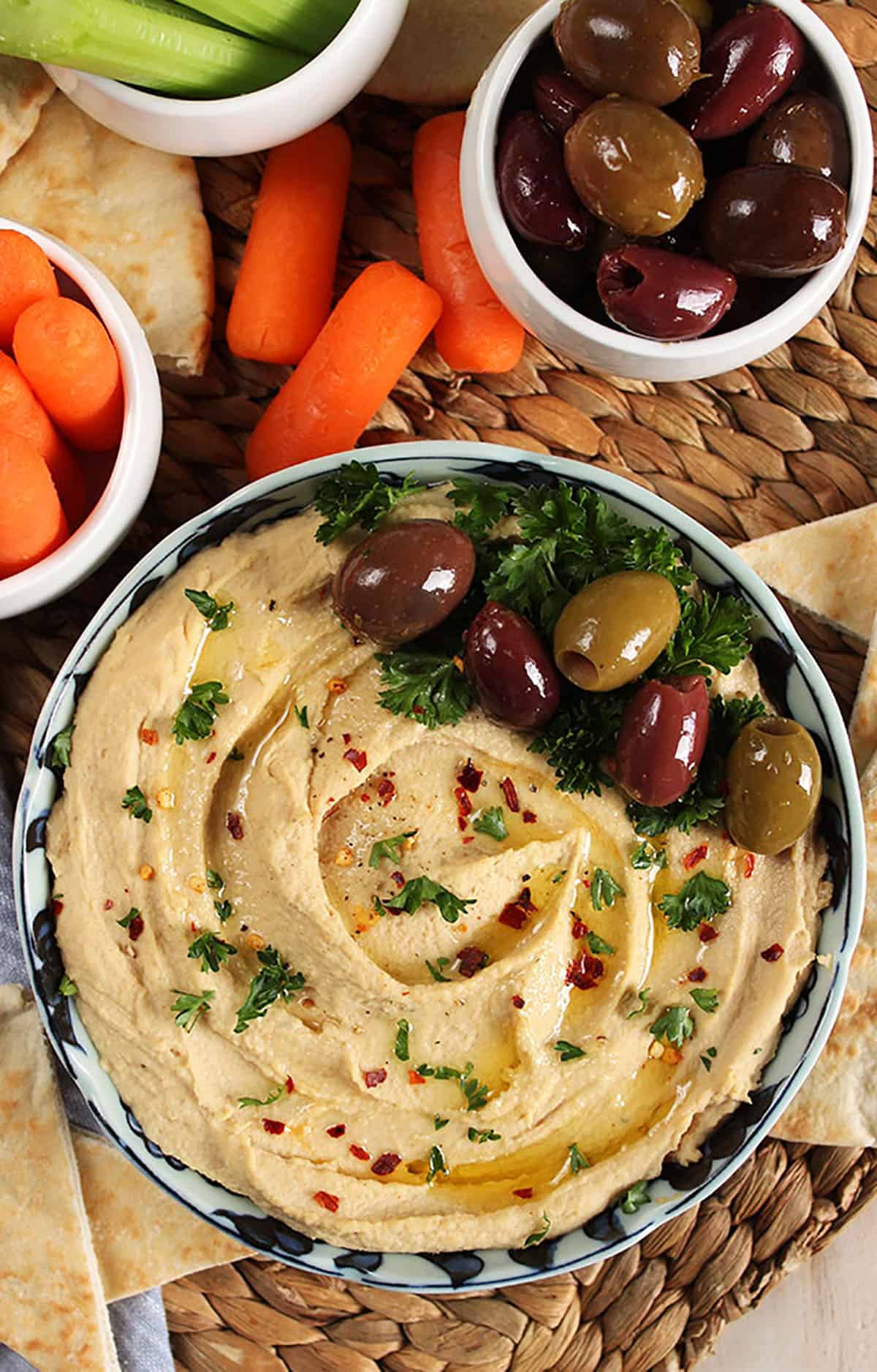 Hummus in a bowl topped with olives on a wicker placemat with pita wedges, carrots and celery for dipping.