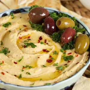 Hummus in a blue and white bowl topped with crushed red pepper, olives, parsley and a drizzle of olive oil with pita wedges around.
