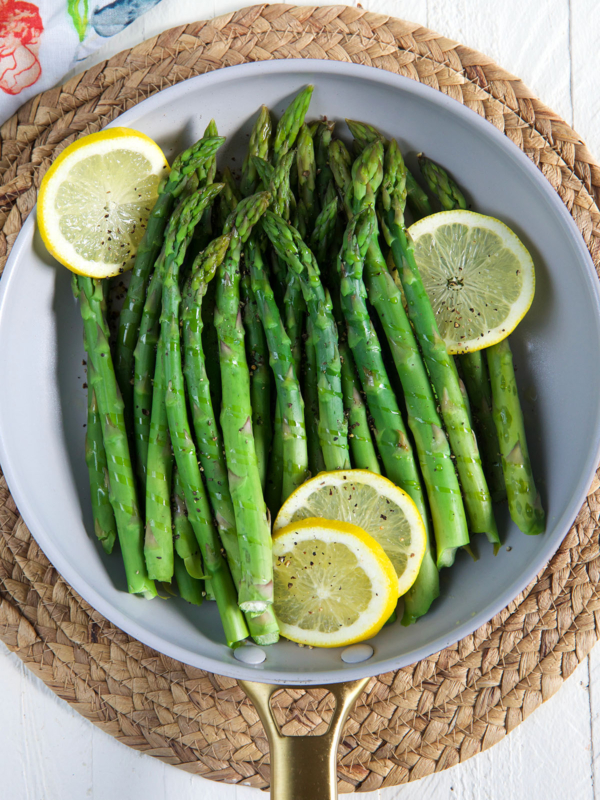 Steamed asparagus is presented in a round skillet with several lemon slices.