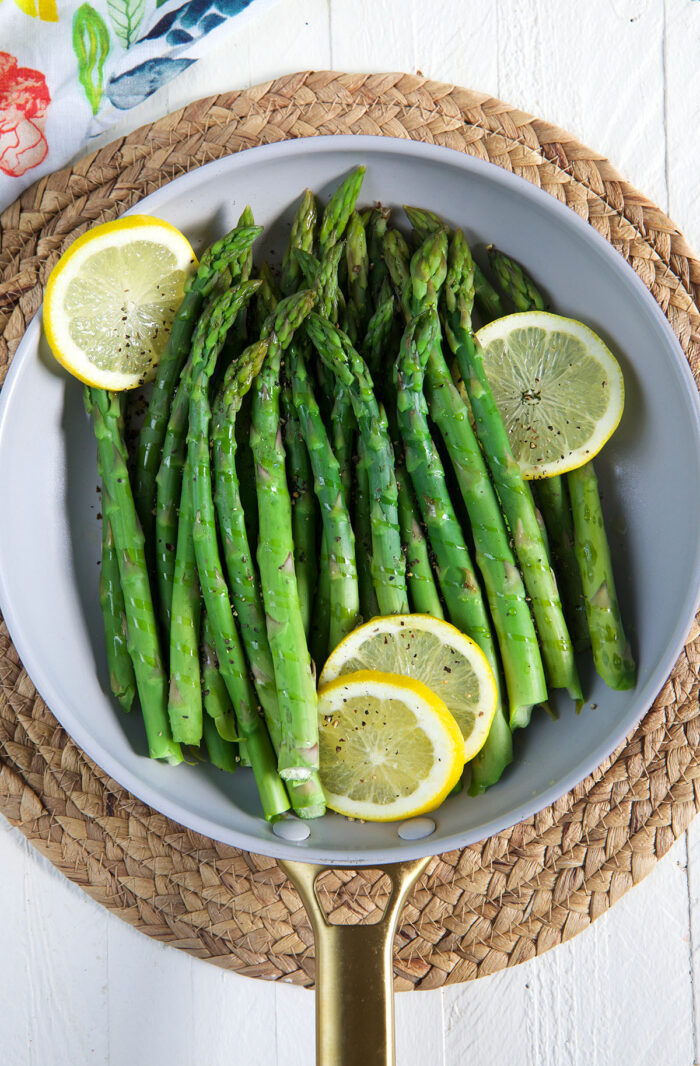 Steamed asparagus is presented in a round skillet with several lemon slices.