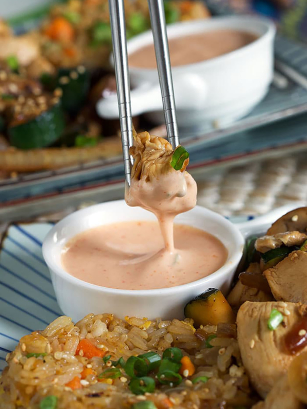 a piece of chicken being dipped into a white bowl with yum yum sauce with stainless steel chopsticks.