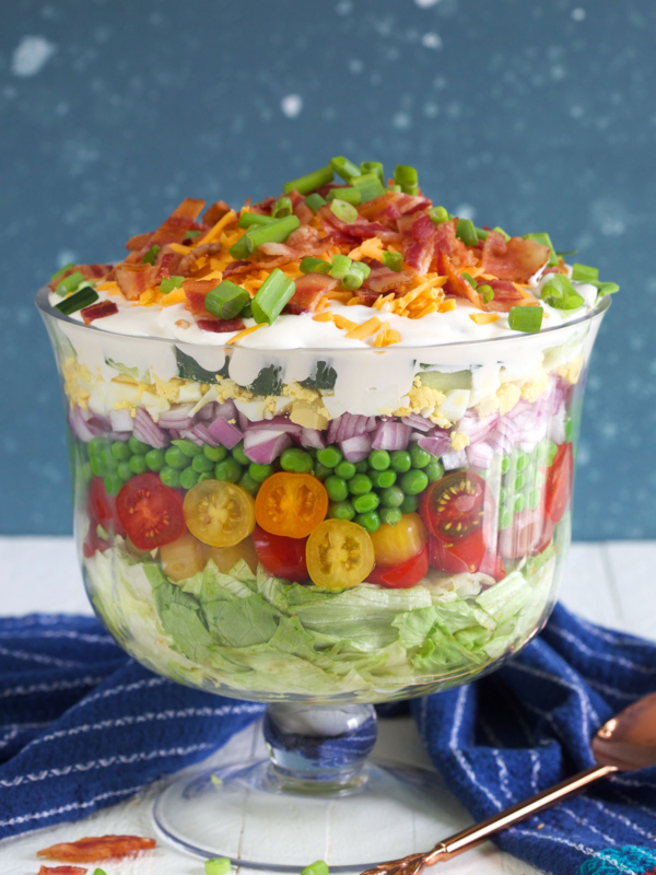 A seven layer salad is prepared in a trifle dish on a white countertop.