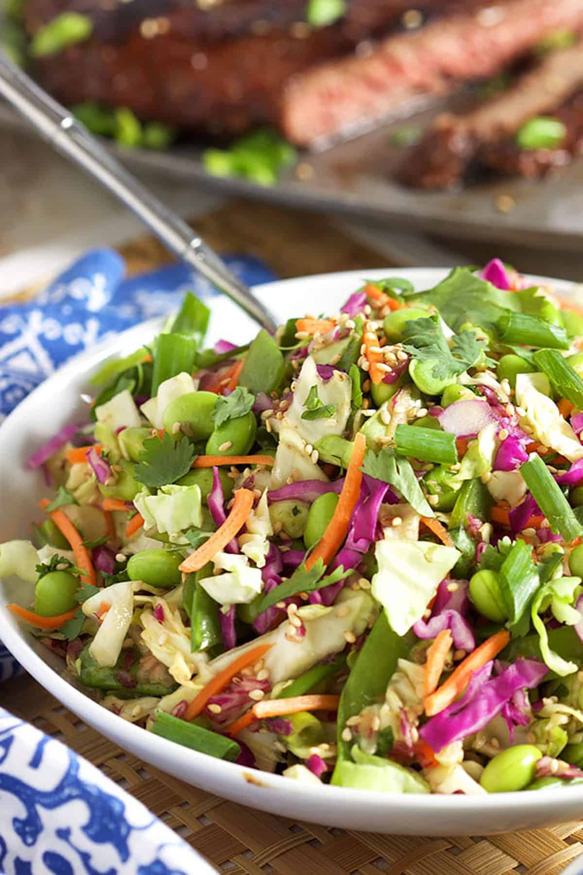 Asian slaw in a white bowl with a silver spoon and a blue and white napkin to the left.