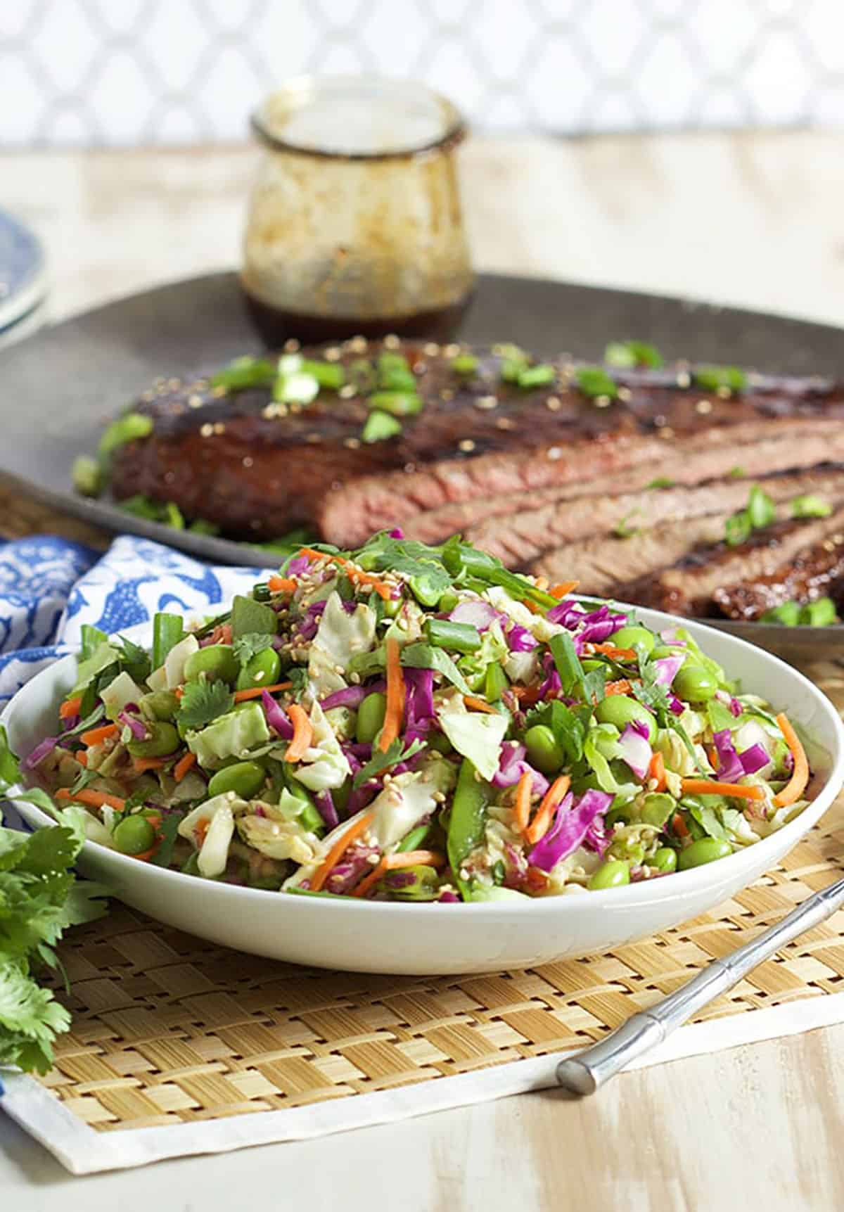 Asian slaw in a white bowl with sliced flank steak in the background on a pewter platter.