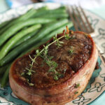 Bacon wrapped filet mignon with thyme sprigs on top and green beans on the plate.
