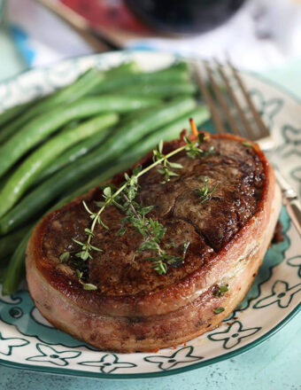 Bacon wrapped filet mignon with thyme sprigs on top and green beans on the plate.