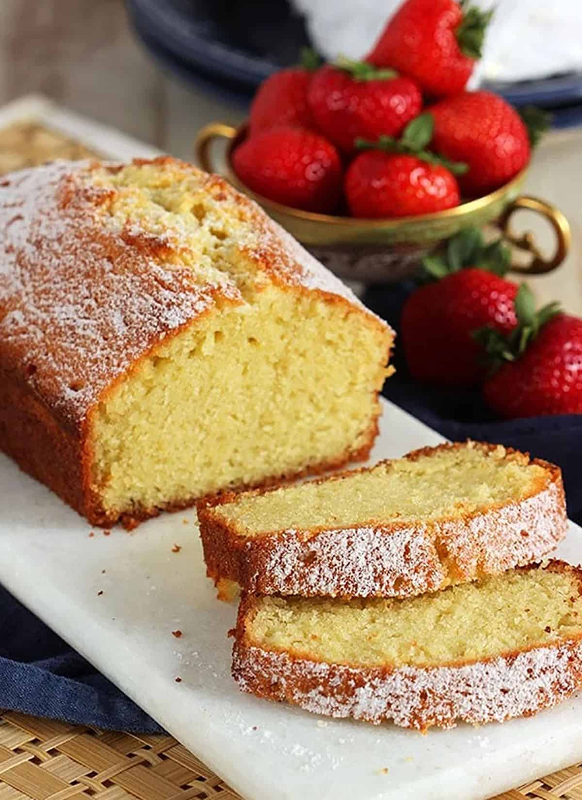 Pound Cake loaf on a white board with two slices stacked next to it and a bowl of strawberries behind it.