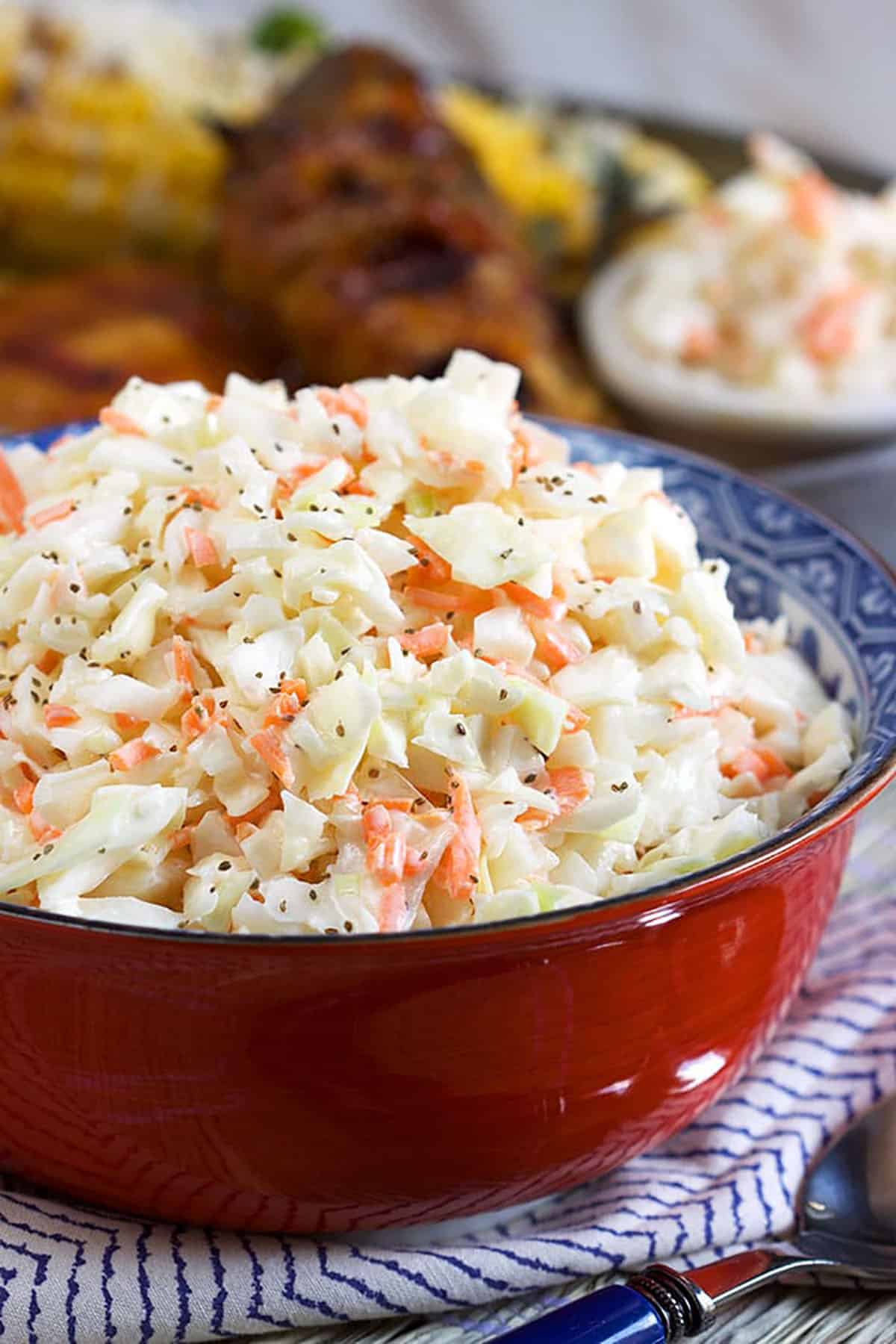 Coleslaw in a red bowl on a blue and white striped napkin with chicken in the background.