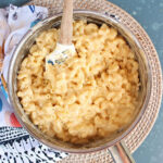 Finished Mac and cheese recipe in a silver saucepan with a wooden spatula on a blue background