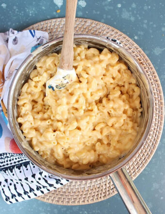 Finished Mac and cheese recipe in a silver saucepan with a wooden spatula on a blue background