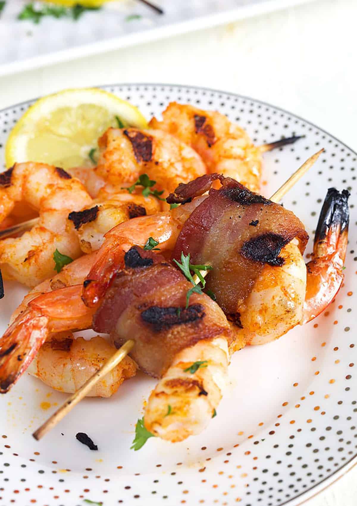 Bacon wrapped shrimp on a bamboo skewer on a white plate with gold polka dots and a lemon slice on the plate.