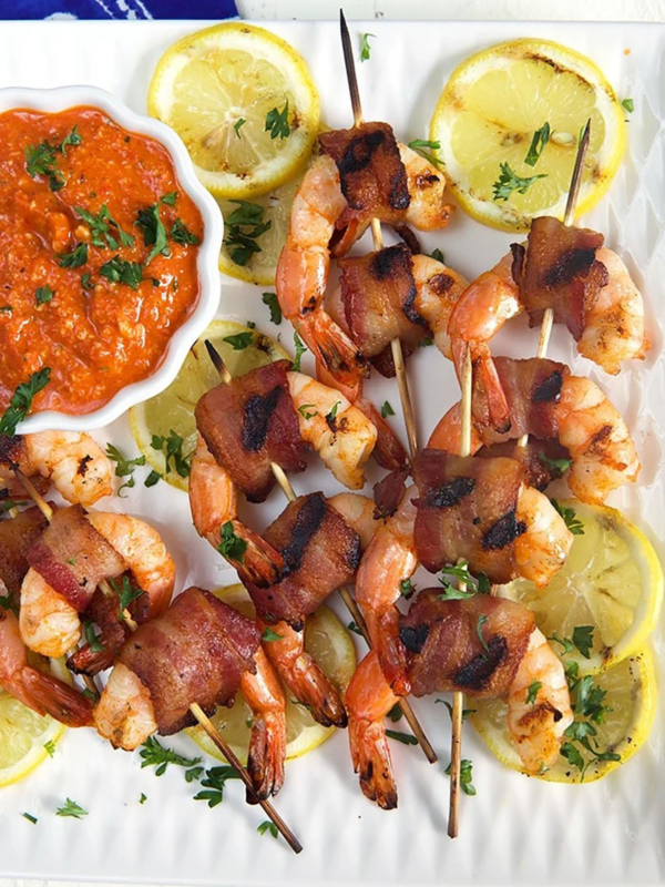 Overhead view of bacon wrapped shrimp on bamboo skewers on a white platter with red romesco sauce in a white bowl.