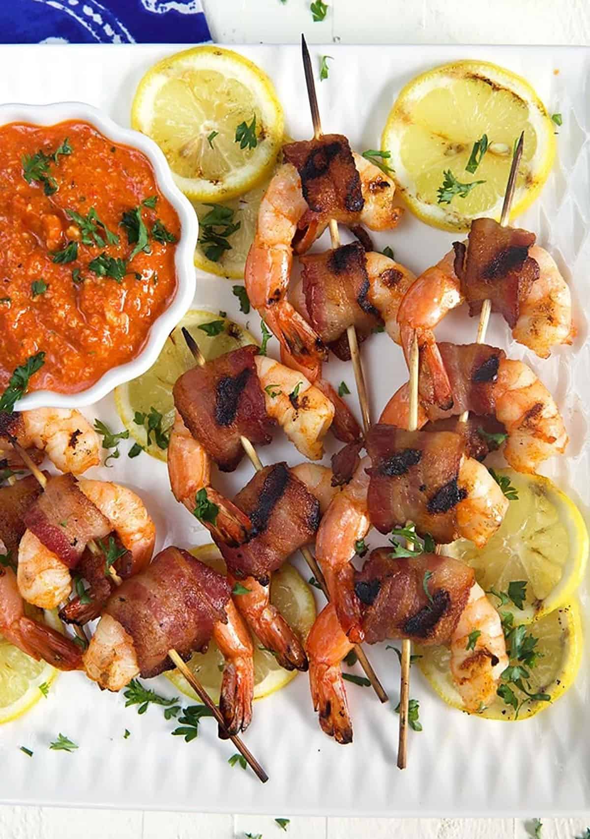 Overhead view of bacon wrapped shrimp on bamboo skewers on a white platter with red romesco sauce in a white bowl.