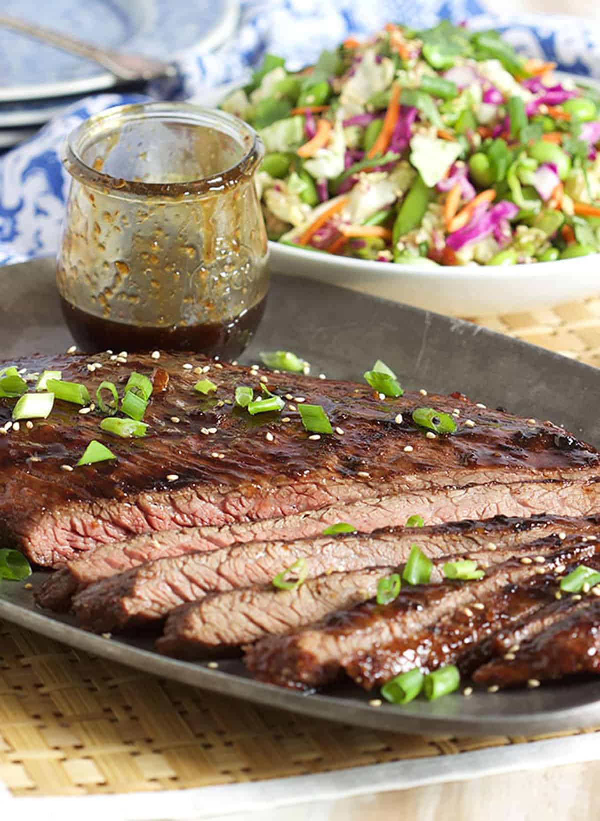 sliced Grilled Flank Steak with teriyaki sauce in a jar on a pewter platter.
