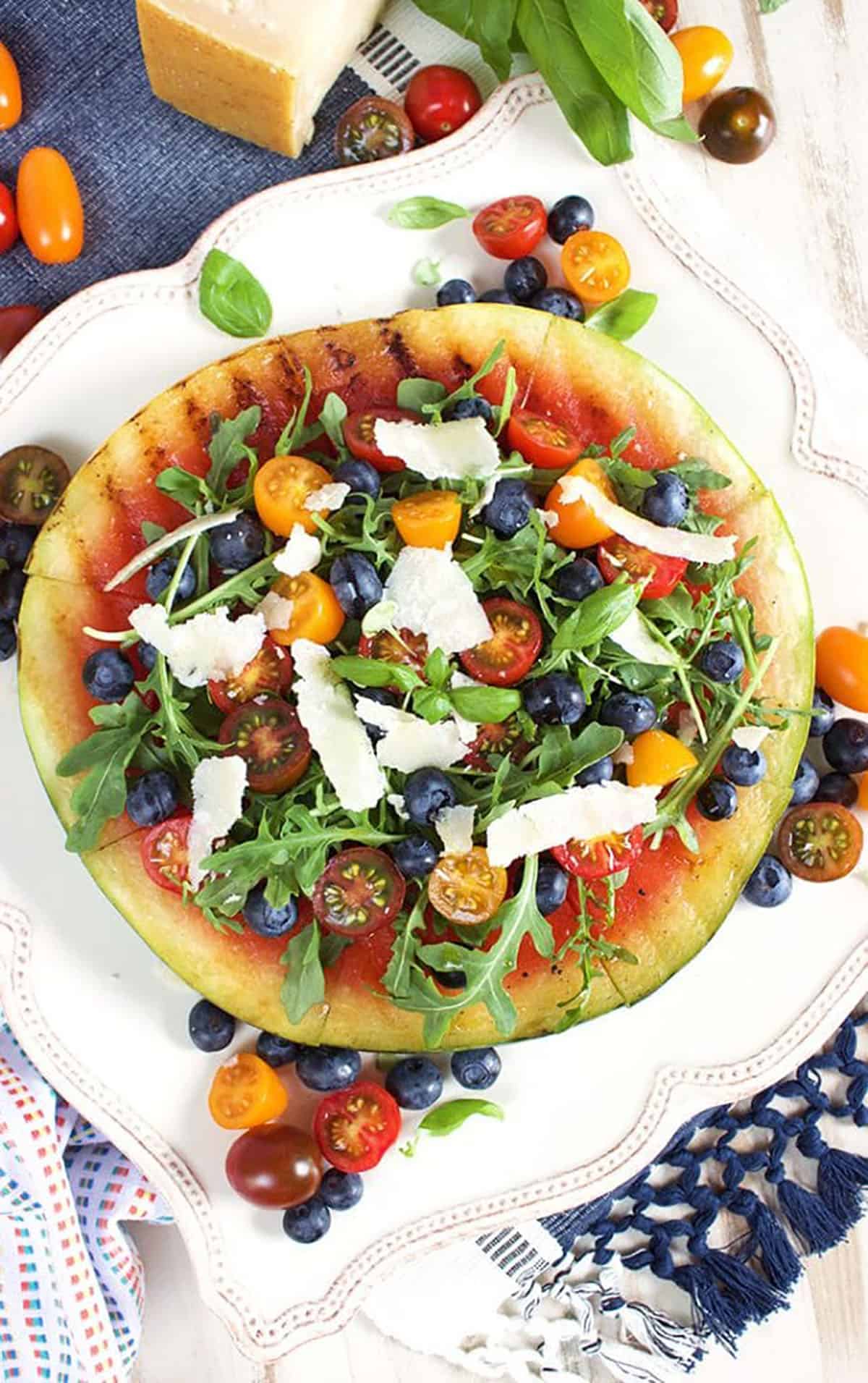 Grilled Watermelon Pizza on a square white platter topped with arugula, berries and parmesan cheese.