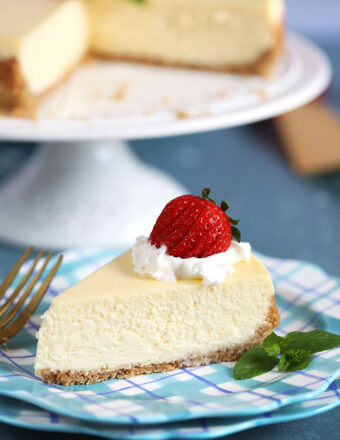 A slice of New York Cheesecake on a blue plaid plate with a strawberry and whipped cream on top and a sprig of mint.
